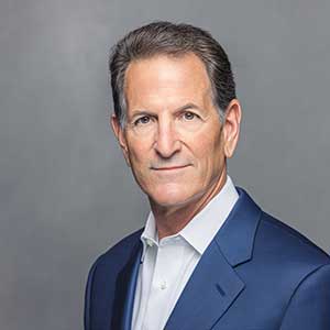 Professional middle-aged man with short dark hair wearing a navy blue suit and white shirt against a gray background.