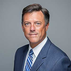 Professional portrait of a middle-aged man in a blue suit and striped tie against a gray background.