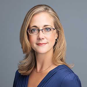 Professional headshot of a woman with shoulder-length blonde hair, wearing glasses and a blue top, against a gray background.
