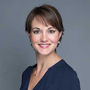 Smiling woman with short brown hair wearing a navy blue top and pearl earrings against a gray background.