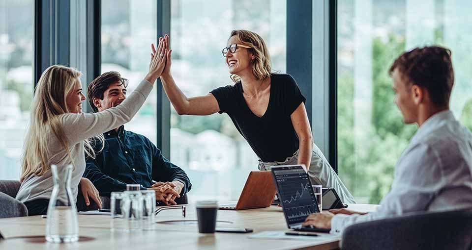 Four colleagues in a modern office celebrating with a high-five around a table with laptops and water glasses.