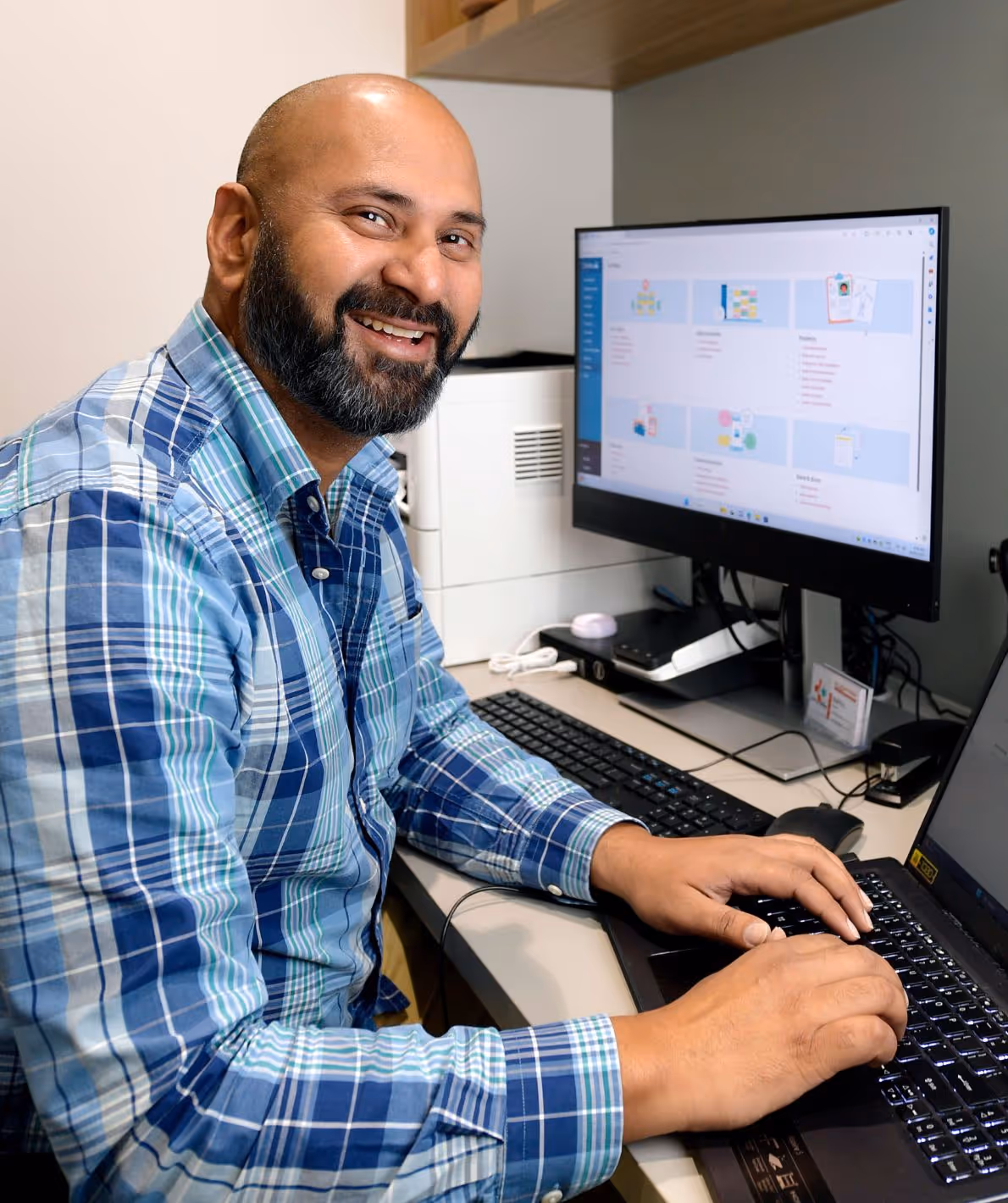 A man with a beard and a blue plaid shirt is smiling while using a computer.