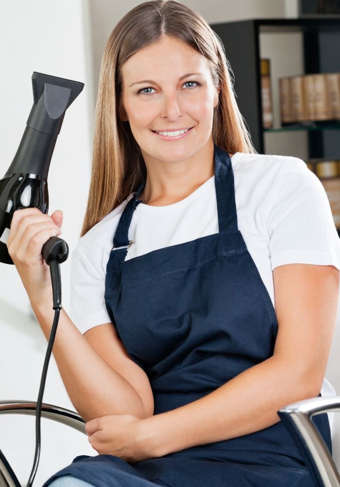 A woman in a white shirt and blue apron holding a hair dryer. 