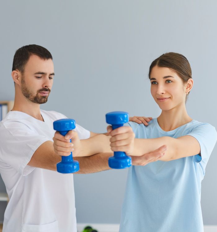 A woman is holding blue weights while a man in white shirt is straightening her pose.