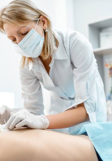 A woman in white coat and wearing a mask is treating a patient's back.