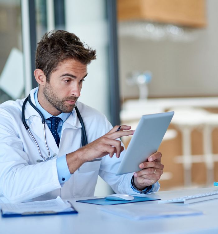A male doctor is focused on a tablet computer, likely reviewing patient information.