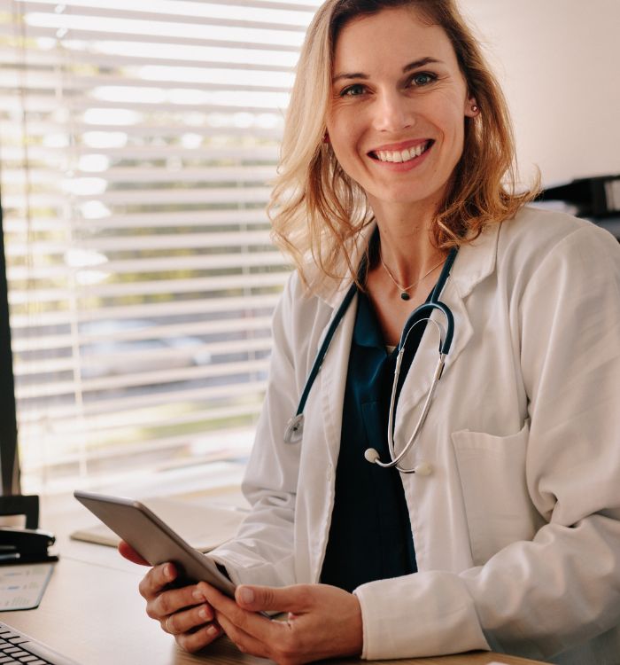 A woman wearing a white coat is seated at her desk, using a tablet for work.