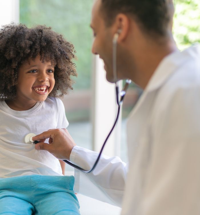 A little girl sitting infront a doctor smiling while the doctor examines her with a stethoscope.
