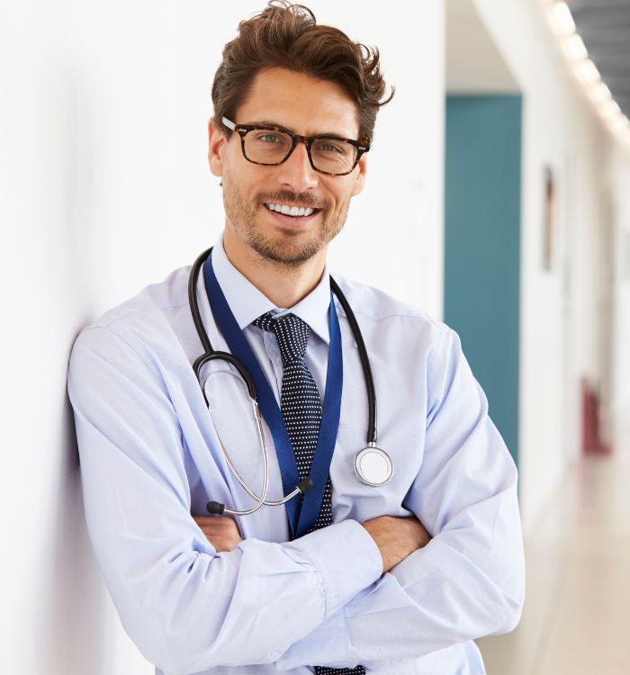 A man wearing a doctor's coat and tie poses in a hospital hallway.