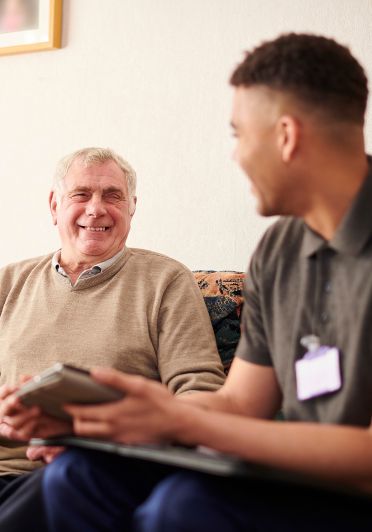 An elderly man and young man sit together on a couch chatting.