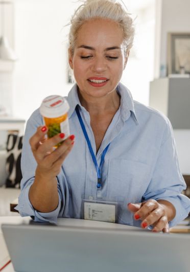 A woman examines a pill bottle in one hand, gazing intently at her laptop, suggesting she is reviewing health information.