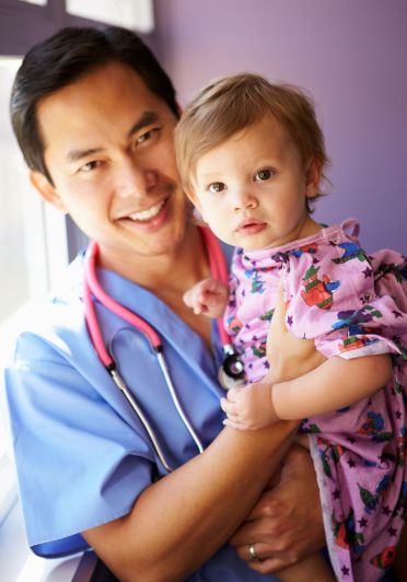 A man wearing a stethoscope holds a baby, smiling gently as he cradles the child in his arms.