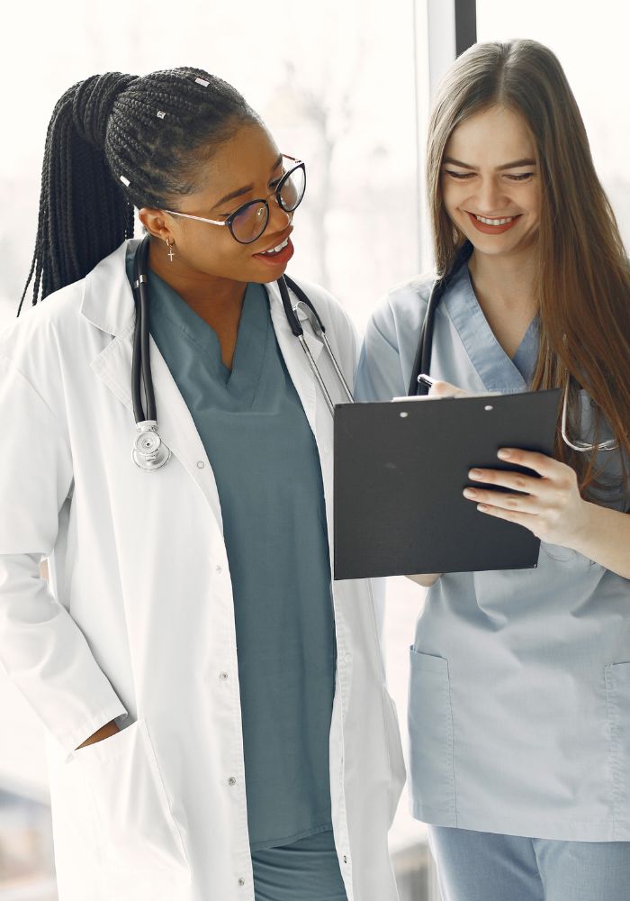 Two female doctors in white coats standing together discussion data on clipboard.