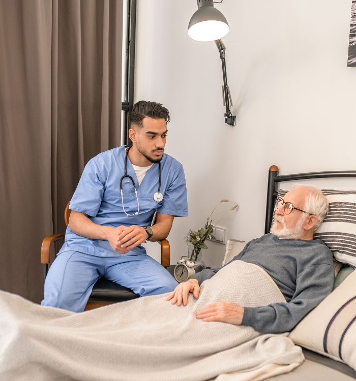 A doctor converses with an elderly man who is resting in bed, providing care and support.