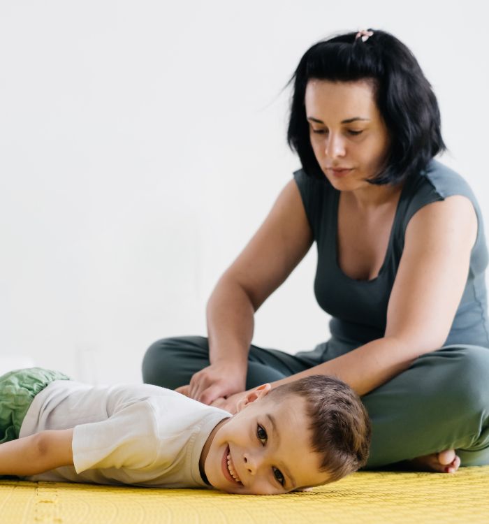 A woman is gentily massaging a child while he is lying comfortably on a vibrant yellow mat.