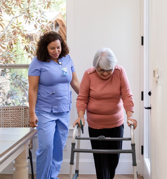 An older woman uses a walker while being supported by a younger woman beside her.
