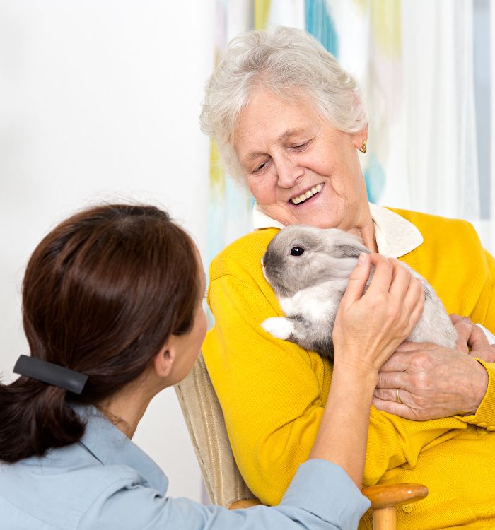 A woman sitting in a chair, gently holding a small rabbit in her hands, smiling softly at the animal.