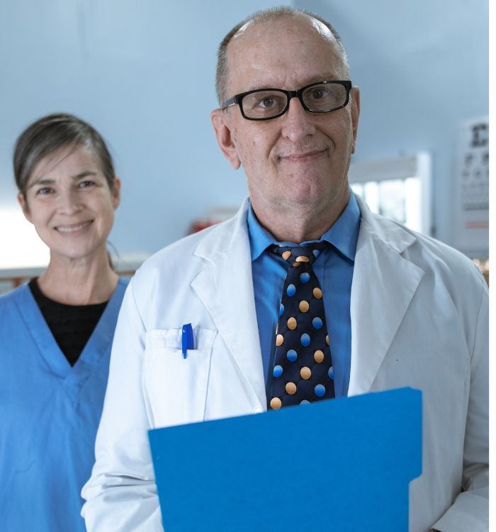 A woman in blue scrub, and a man in a lab coats and holding blue folder are standing together