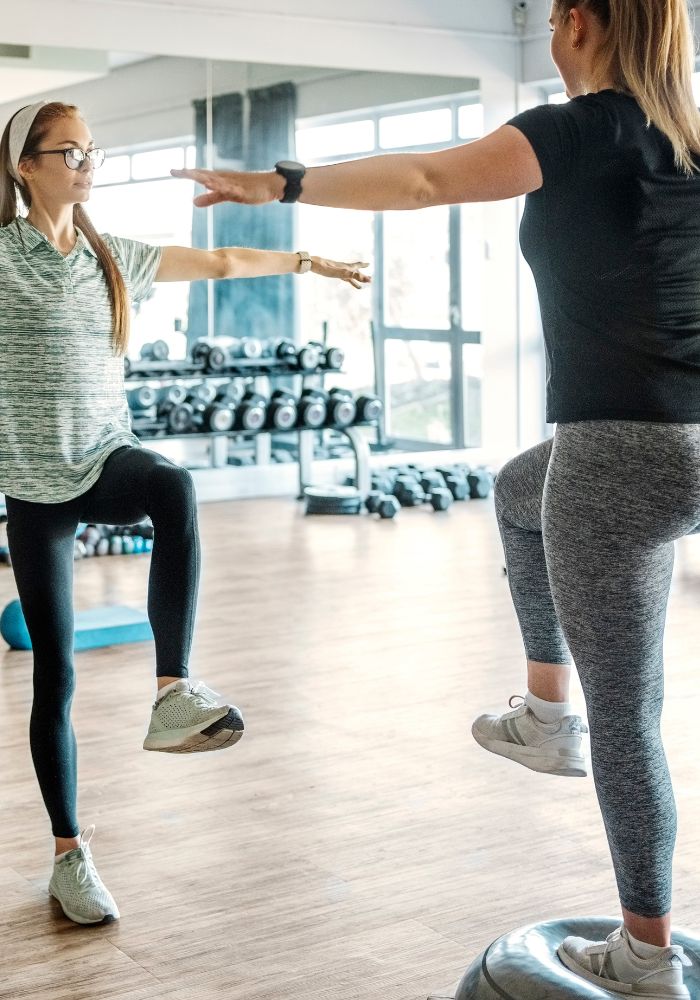 Two women are in a gym, one is jumping and the other is holding her hands out.