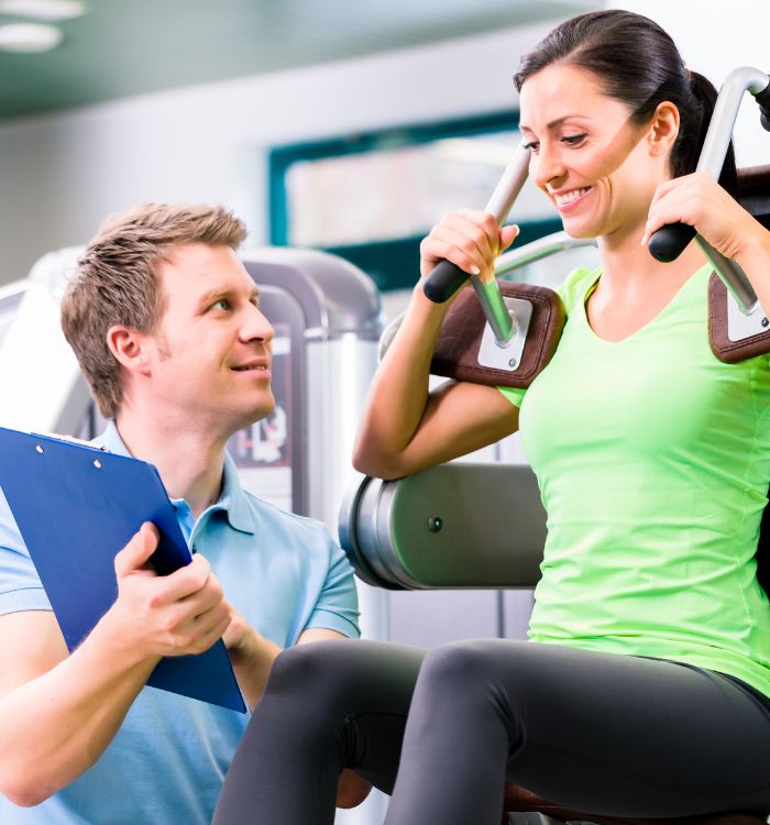 A man and woman are in a gym, the woman is on a machine and the man is holding a clipboard.