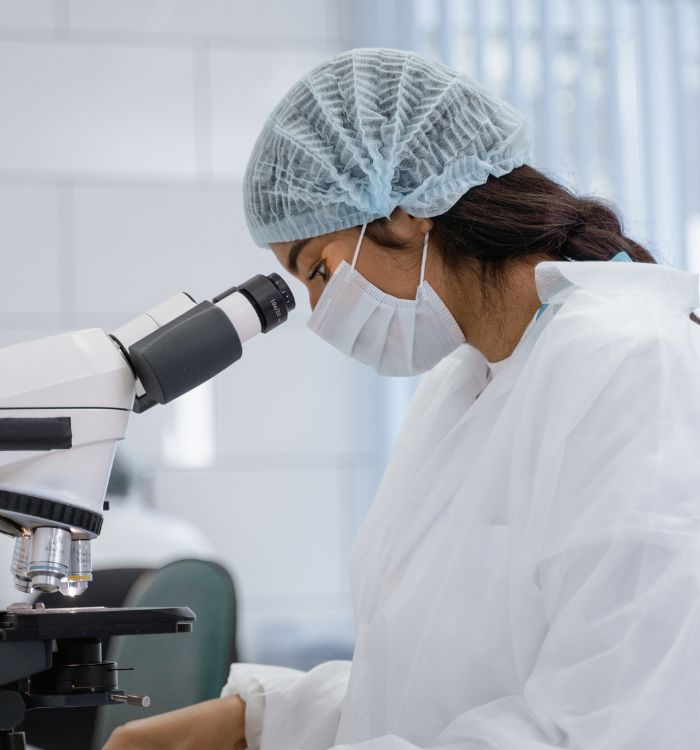 A woman wearing a white lab coat and a blue hat looking through a microscope.