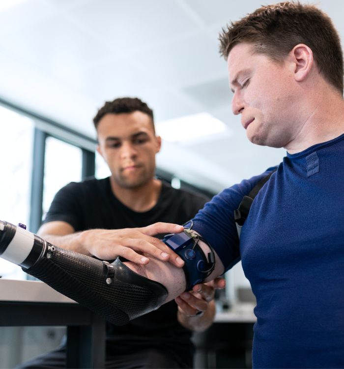 A man with a blue shirt and black pants is adjusting a brace on another man's arm.