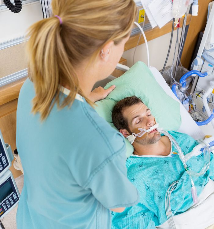 A woman in a blue shirt is helping a man in a hospital bed.