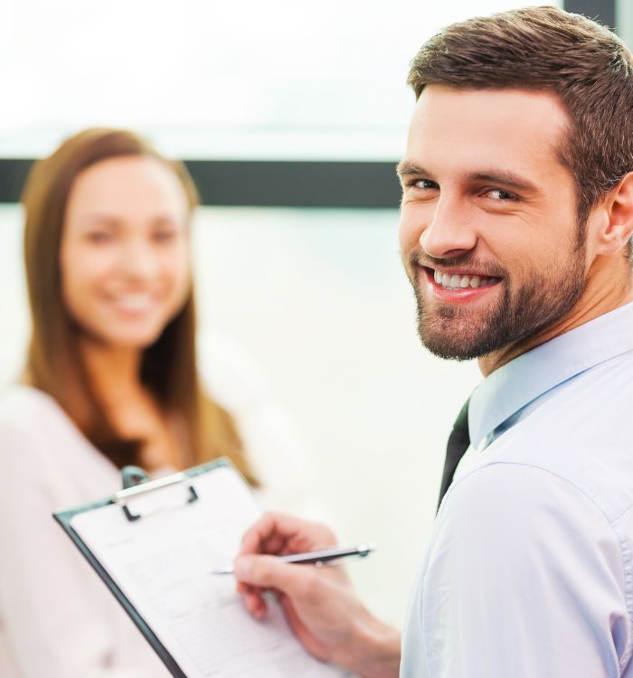 A man in a white shirt is smiling while writing on a clipboard.