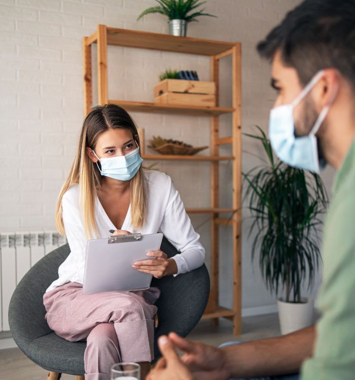 A woman wearing a face mask sits in a chair and talks to a man.