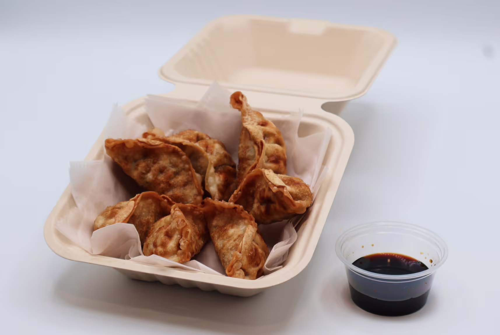 Takeout container with fried dumplings and a small cup of dark dipping sauce on a white surface.