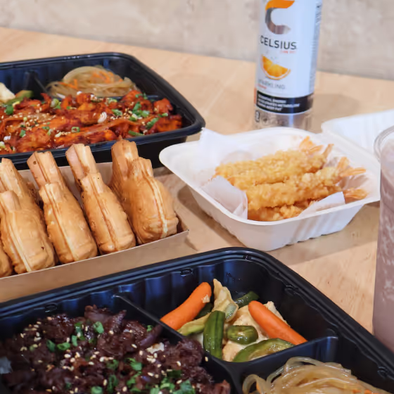 Assorted Asian meal with fried shrimp, grilled meat, vegetables, sandwiches, and a drink on a wooden table.