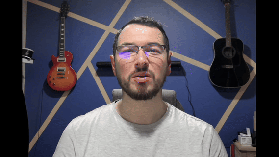 Man with glasses and a beard speaking in front of a blue wall with geometric wood strips and two guitars hanging behind him.
