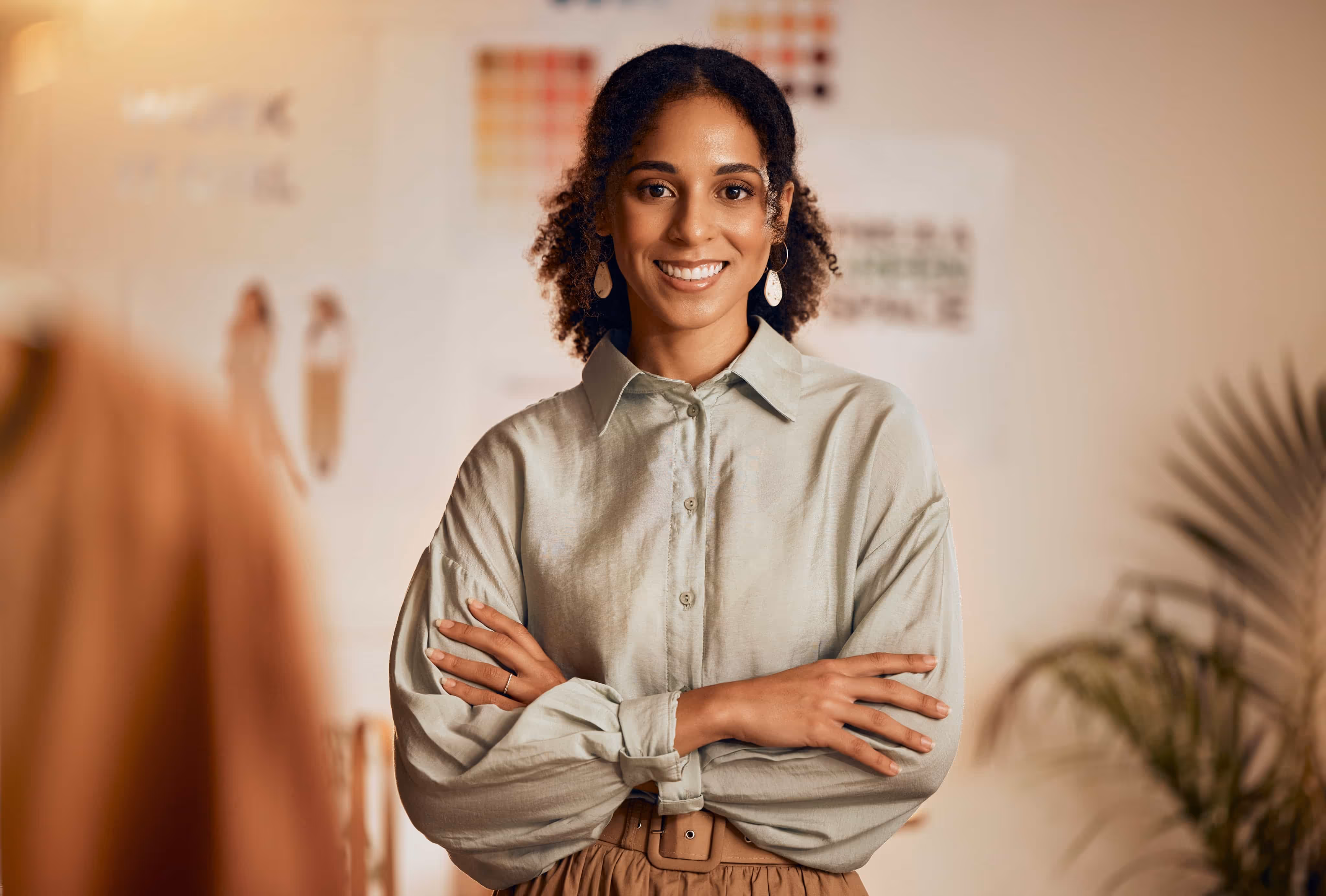 Smiling woman with curly hair wearing a light shirt, standing with arms crossed indoors.