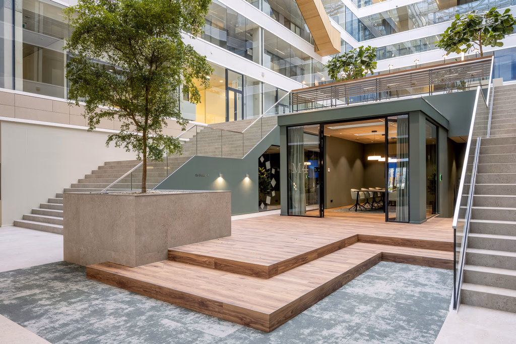 Modern office atrium with wooden steps, a large planter with a tree, glass-enclosed meeting room, and staircases on both sides.