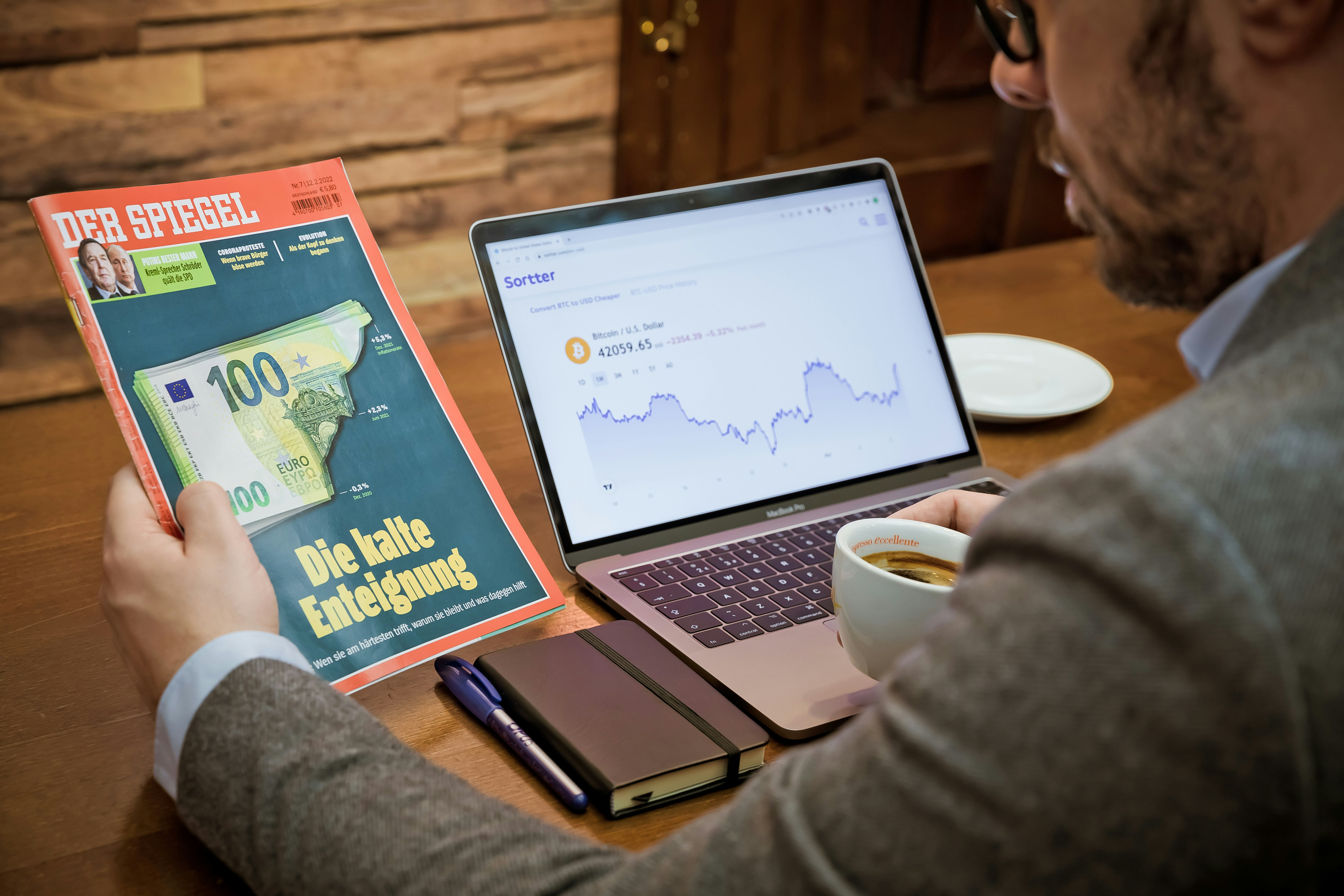 Man holding a Der Spiegel magazine showing a folded 100 euro bill with text about expropriation, sitting at a table with a laptop displaying Bitcoin price chart and holding a cup of coffee.