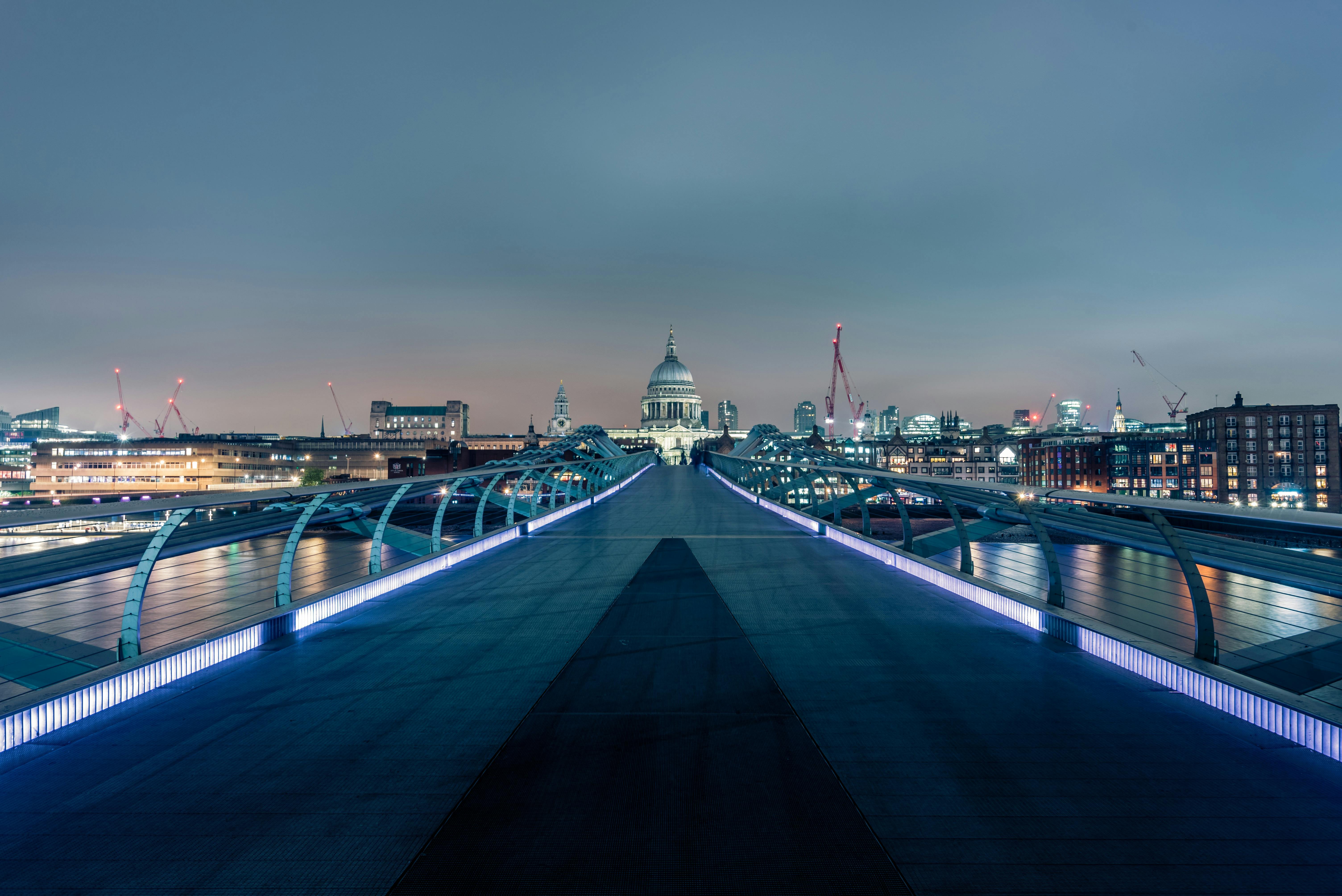 Night view of Millennium Bridge leading towards illuminated St. Paul's Cathedral in London with city buildings and cranes in the background.