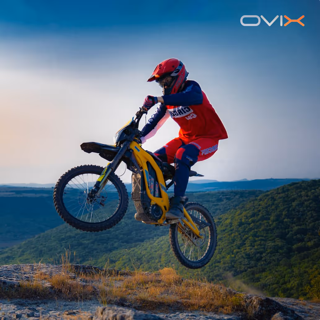 Motorcyclist wearing red gear jumping a yellow dirt bike over rocky terrain with hills in the background.