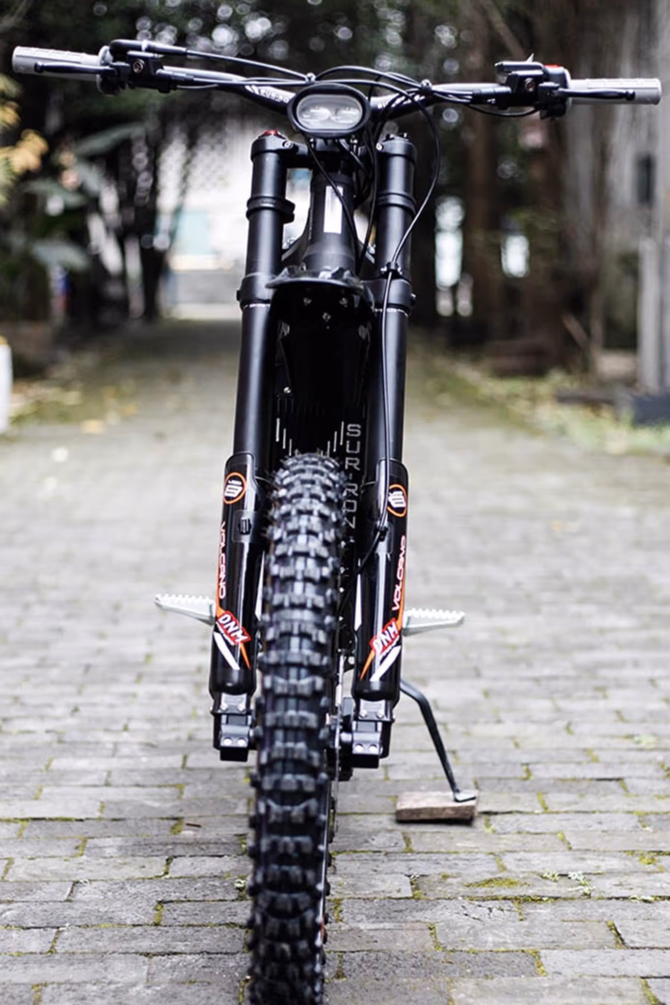Front view of black Sur-Ron electric dirt bike with knobby tire on stone pathway.