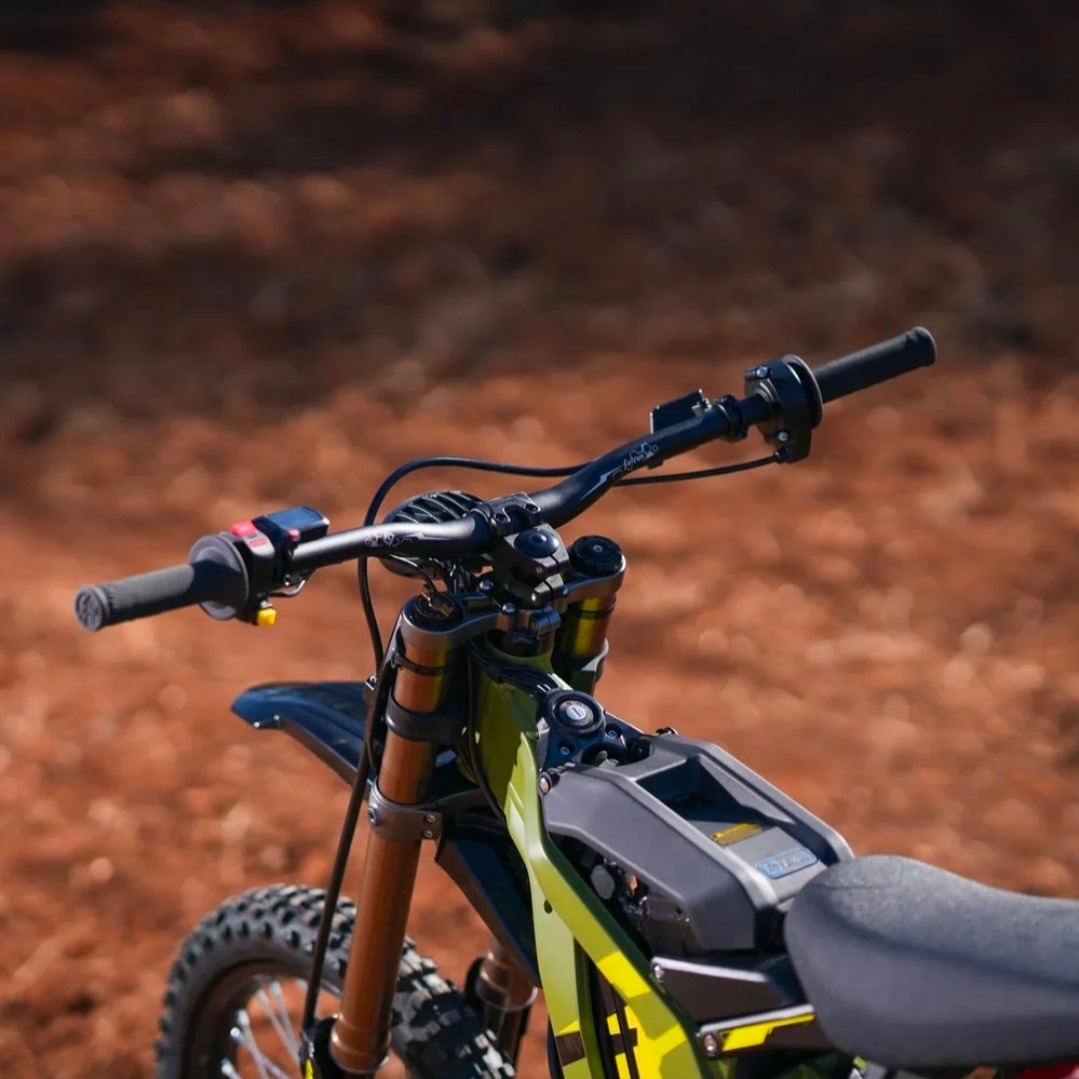 Close-up of green electric dirt bike handlebars and front suspension on a dirt track.
