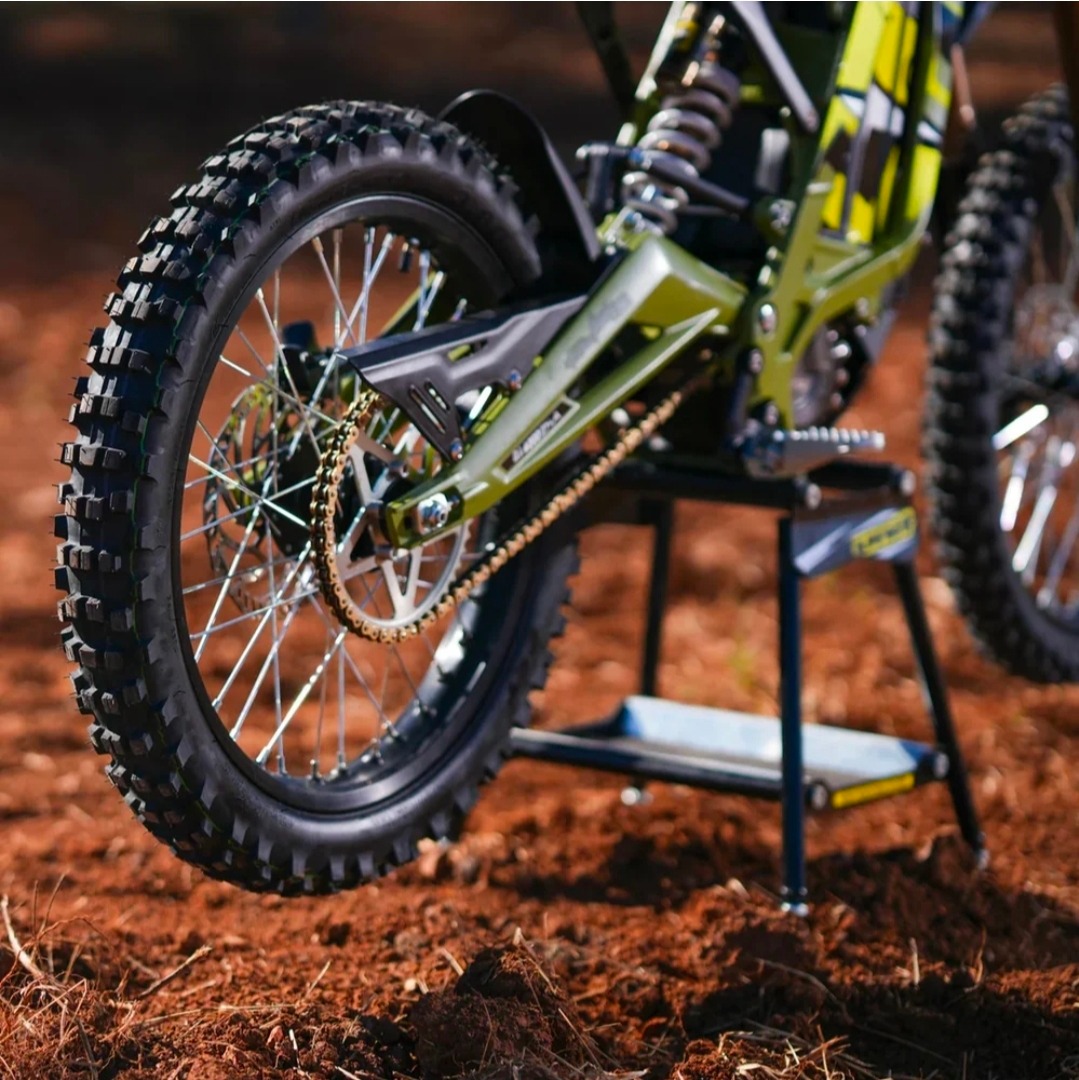 Close-up of the rear wheel and chain of a green dirt bike on reddish soil.
