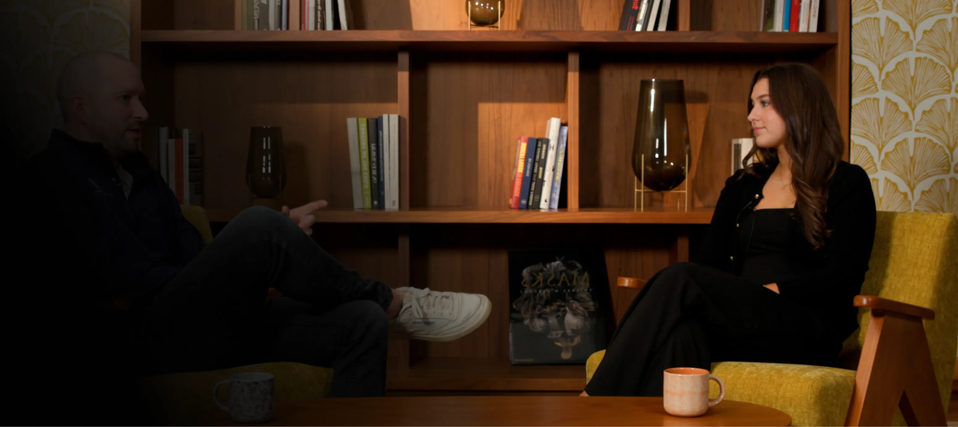 Two people seated in a cozy room with wooden shelves, having a conversation across a table with mugs.