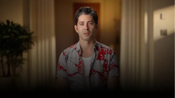 Man with dark hair wearing a white and red patterned shirt sitting indoors with blurred background.