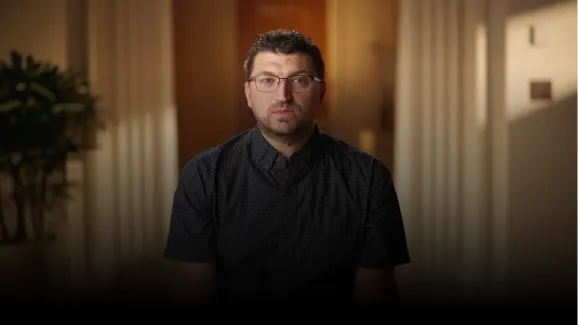 Man with glasses and short curly hair sitting indoors in a softly lit room with a plant in the background.