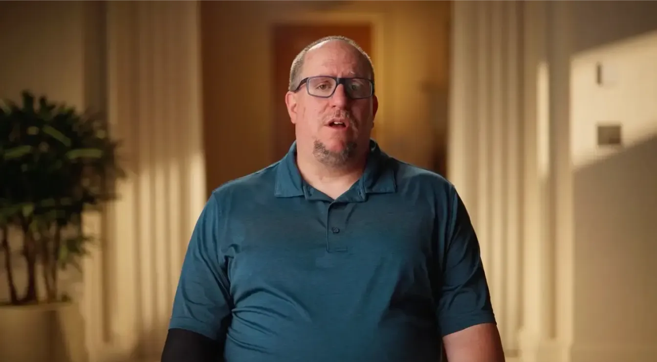 Middle-aged man with glasses and a blue polo shirt sitting indoors with blurred background of columns and a plant.