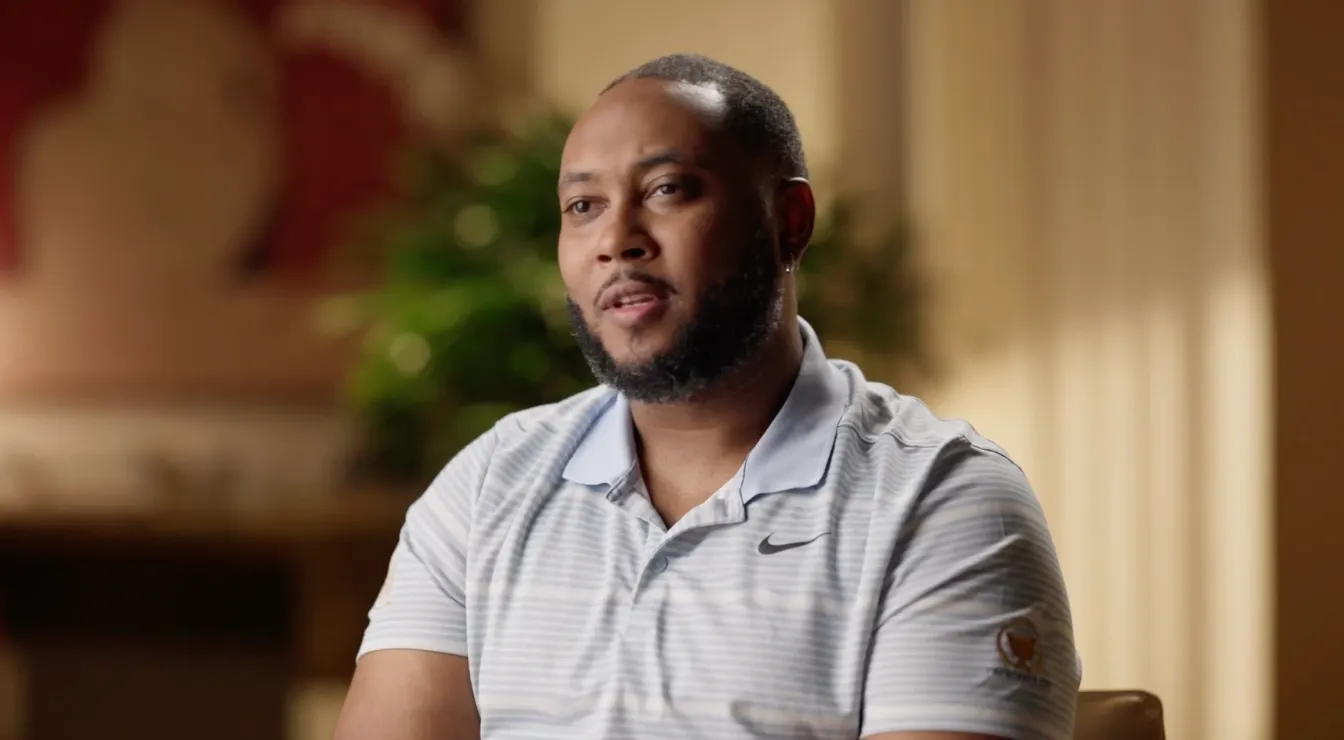 Man with a beard wearing a light gray striped Nike polo shirt sitting indoors with blurred background.