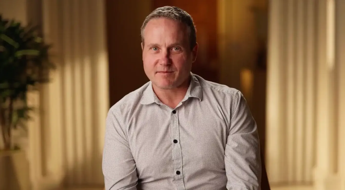 Middle-aged man with short hair wearing a light-colored checkered shirt sitting indoors with blurred curtains and plant in the background.