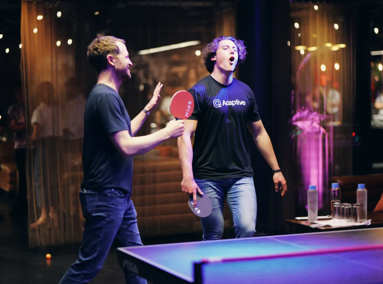 Two men playing ping pong indoors with paddles labeled SPIN, one man in an Adaptive t-shirt showing excitement.