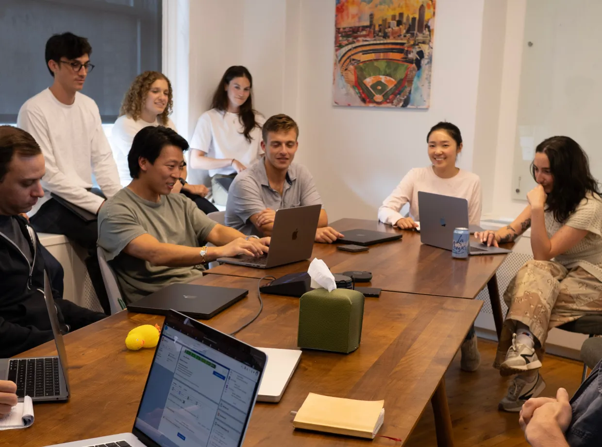 Group of diverse young people sitting around a wooden table in a casual meeting room using laptops and smiling.