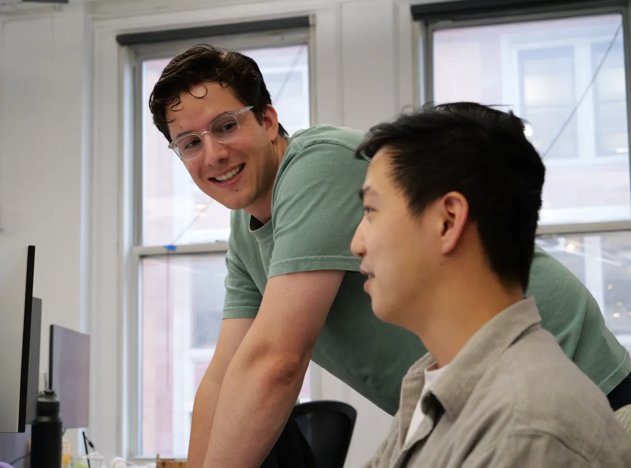 Two men collaborating at a desk with a computer in a bright office with large windows.