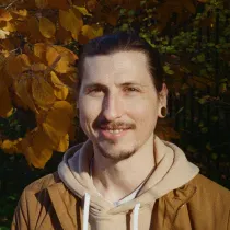 Smiling man with dark hair tied back, wearing a brown jacket and beige hoodie, standing outdoors with autumn leaves in the background.